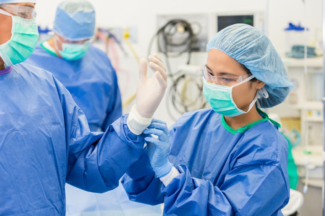 Surgeon wearing mask, protective eyewear, hair net, gown, and helping other surgeon with sterile gloves before operation.
