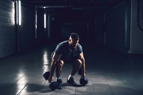Close up view of focussed hardworking active fitness strong muscular bearded bodybuilder man crouching before raising heavyweight dumbbell in the underground garage or urban gym while looking far away.